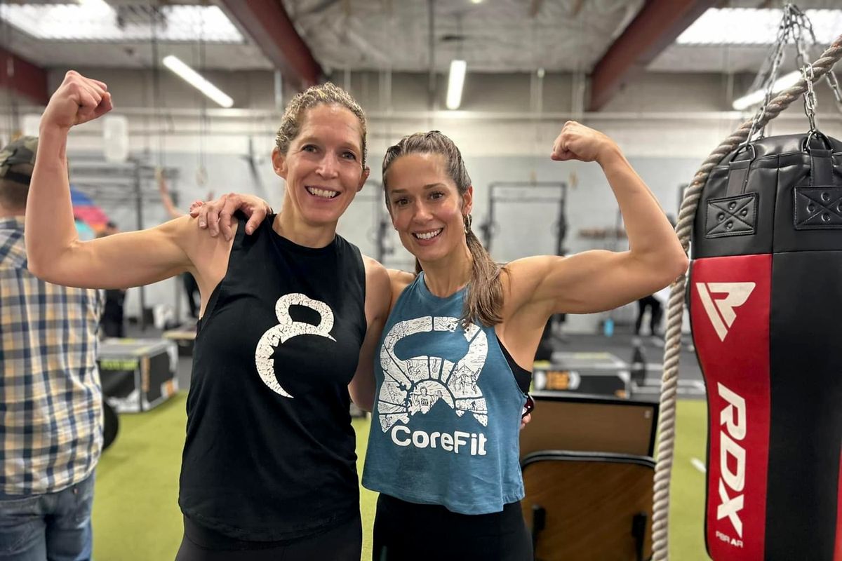 A CoreFit member stands shoulder to shoulder with trainer Betsy, both flexing their arm muscles.