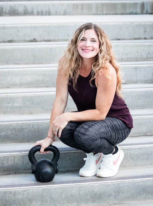 Lacy crouches on a flight of stairs with a kettlebell in hand and a smile on her face.