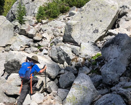 A child with a backpack and walking poles hikes up a steep incline with many large rocks. His boot rests on the next rock, ready to take a step. This is going to be easy after all that coaching, he is thinking.