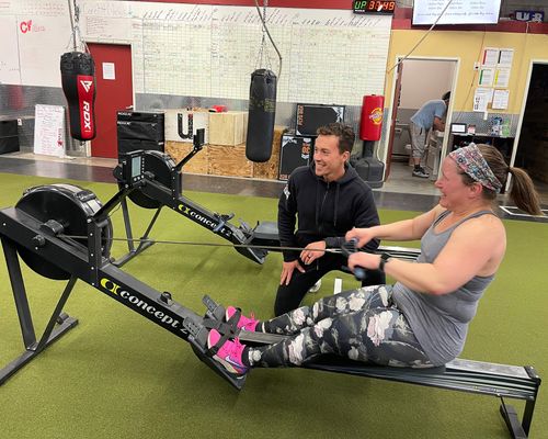 Owner Kurt kneels beside and encourages a CoreFit member on a rowing machine, both clearly enjoying the moment.