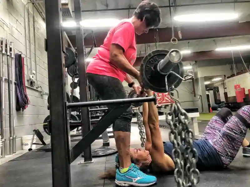 One CoreFit member spots another who is laying on the ground doing overhead presses with a barbell.
