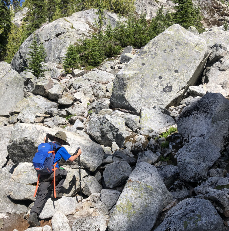 A child with a backpack and walking poles hikes up a steep incline with many large rocks. His boot rests on the next rock, ready to take a step. This is going to be easy after all that coaching, he is thinking.