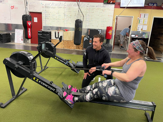 Owner Kurt kneels beside and encourages a CoreFit member on a rowing machine, both clearly enjoying the moment.