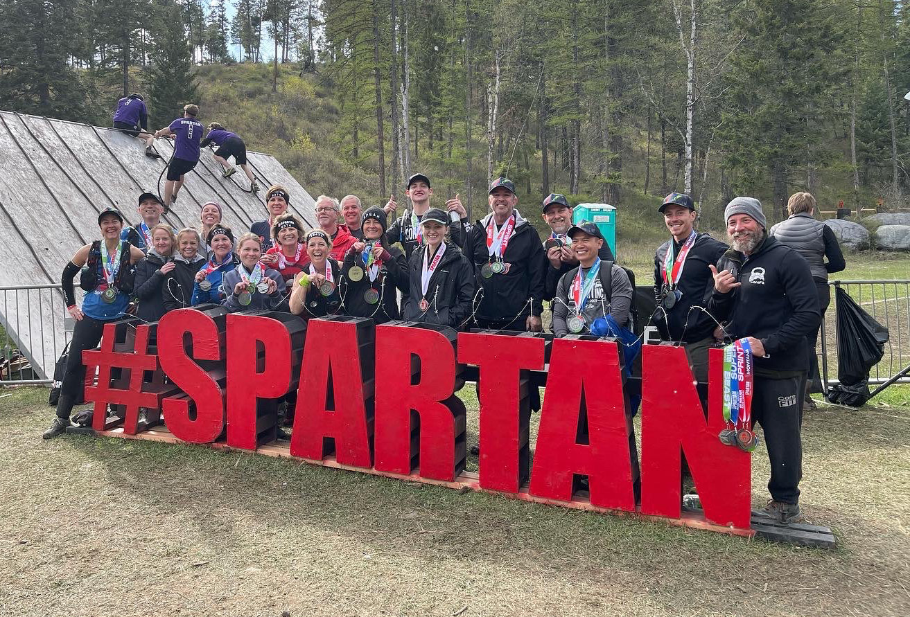 A CoreFit crew of about 20 people hold their finishing medals while standing behind a large sign that says #SPARTAN.
