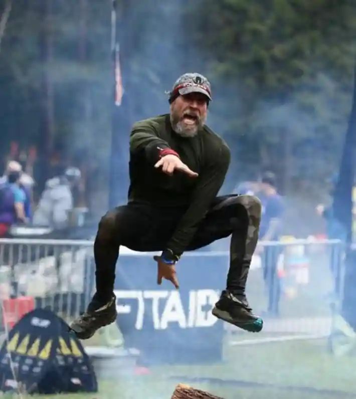 Darren leaps over flames and smoke at the finish line of a Spartan Race, knees out to the sides and fingers forming heavy metal horns, with one arm straight out in front of him and the other toward the ground. His eyes are obscured by the bill of his hat, but his mouth is open in the universal expression of 'victory'.